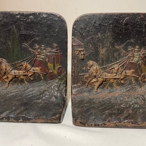 May include: A pair of antique cast iron bookends featuring a horse-drawn carriage with three horses and three passengers. The bookends are painted in shades of black, brown, gold, and white.