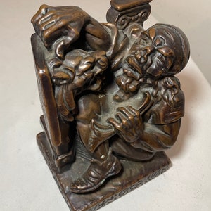 Puede incluir: Una escultura de bronce de un hombre con barba sentado en un banco. El hombre sostiene un libro en su mano izquierda y un rollo en su mano derecha. La escultura es detallada y realista.