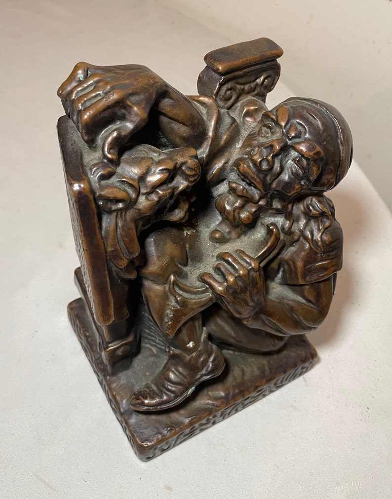 Puede incluir: Una escultura de bronce de un hombre con barba sentado en un banco. El hombre sostiene un libro en su mano izquierda y un rollo en su mano derecha. La escultura es detallada y realista.