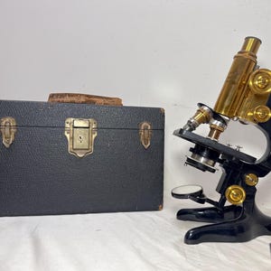 May include: An antique microscope with brass and black components, next to a black case. The microscope has a gold-colored tube and adjustment knobs. The case has a textured surface and a metal clasp.