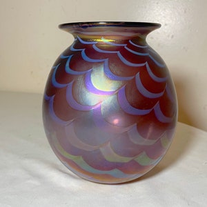 May include: A small, round iridescent glass vase with a scalloped pattern. The vase is a deep purple color with hints of blue and gold.