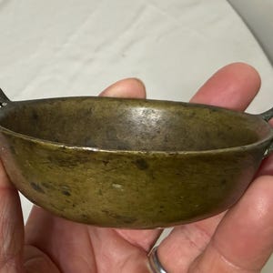 Puede incluir: Un peque&ntilde;o cuenco de bronce antiguo con dos asas de bucle. El cuenco tiene una forma redondeada y una p&aacute;tina oscura y envejecida. Las asas est&aacute;n unidas a los lados del cuenco, y el dise&ntilde;o general es simple y funcional.