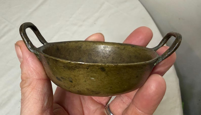 Puede incluir: Un peque&ntilde;o cuenco de bronce antiguo con dos asas de bucle. El cuenco tiene una forma redondeada y una p&aacute;tina oscura y envejecida. Las asas est&aacute;n unidas a los lados del cuenco, y el dise&ntilde;o general es simple y funcional.