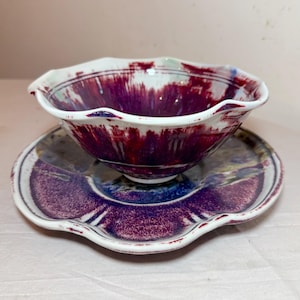 May include: A handmade ceramic bowl and saucer set. The bowl has a ruffled edge and is primarily white with streaks of deep red and purple. The saucer is a matching color scheme with a wavy edge. The set is designed for serving food.