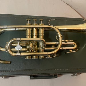 May include: A gold-colored cornet with three valves and a bell, in its black case. The cornet is in good condition and appears to be vintage.