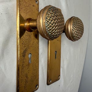 May include: Antique brass door hardware set featuring two rectangular backplates with keyholes and matching woven-patterned doorknobs. The set has a weathered, aged appearance, with visible signs of wear and patina.