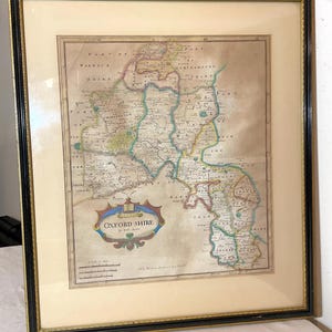 May include: An antique map of Oxfordshire, England, printed in black, brown, green, and blue ink on cream paper. The map is framed in a black and gold frame. The title of the map is "Oxford Shire".