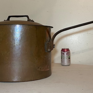 May include: Large antique copper pot with a long handle and a lid. The pot is brown and has a shiny finish. The handle is made of metal and is attached to the side of the pot.