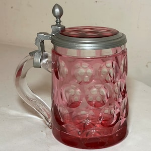 May include: A vintage glass beer stein with a pewter lid and a clear glass handle. The stein is decorated with a pink, bubble-like pattern.