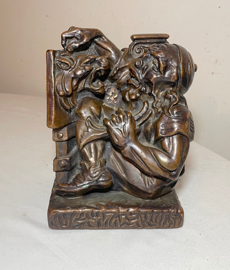 Puede incluir: Un sujetalibros de bronce que representa a un hombre luchando contra una bestia m&iacute;tica. El hombre lleva un casco y una t&uacute;nica, y la bestia tiene una cara aullante y garras. El sujetalibros es detallado y tiene una base texturizada.