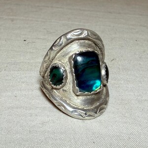 May include: A silver ring with three inlaid stones. The largest stone is rectangular and iridescent with blue and green hues. Two smaller, round green stones flank the larger stone. The ring has a decorative, engraved design.