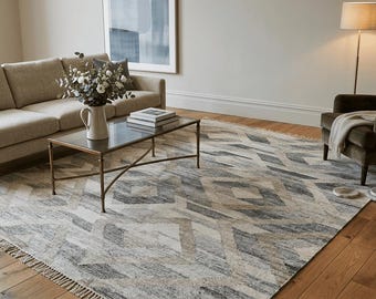 May include: A living room scene featuring a beige sofa, a dark armchair, and a patterned rug. The rug has a geometric design in shades of gray and beige. A glass-topped coffee table and a floor lamp complete the cozy interior.