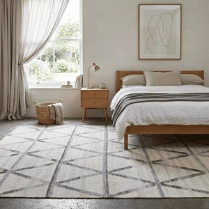 May include: A bedroom interior featuring a wooden bed frame with white bedding and a gray blanket. A geometric patterned rug in gray and white covers the floor. A wooden clothing rack and a small nightstand are also visible.