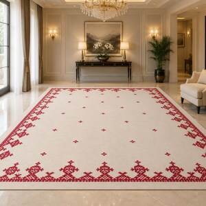 May include: A large rectangular area rug featuring a cream-colored base with a vibrant red geometric border pattern. The design includes intricate cross-stitch style motifs along the edges and small, evenly spaced red accents across the center.