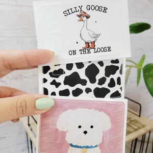 May include: Three white cards with different designs. The top card has a cartoon goose wearing a cowboy hat and boots with the text "Silly Goose on the Loose." The middle card has a black and white cow print. The bottom card has a white dog with a blue collar.