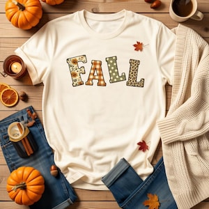 May include: A cream-coloured short-sleeve t-shirt featuring the word FALL printed in large block letters. Each letter has a distinct pattern: sunflowers, orange and white plaid, green with white polka dots, and a brown leopard print design.