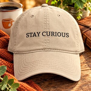 May include: A beige baseball cap featuring the embroidered black text STAY CURIOUS centered on the front panel. The cap has a structured crown, a curved brim with visible stitching, and a button at the top center of the fabric dome.