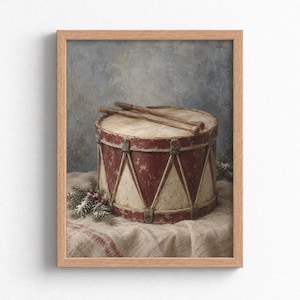 May include: A framed wall art print featuring a weathered red and cream-colored drum with two wooden drumsticks resting on top. The drum is decorated with a triangular pattern and sits on a textured fabric surface next to a small pine sprig.