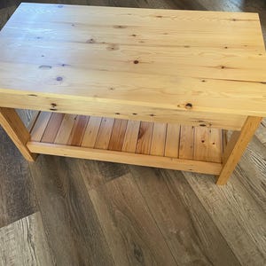 May include: A rectangular coffee table constructed from light-colored natural wood with visible grain patterns and dark knots. The table features a solid flat top surface and a lower slatted shelf supported by four sturdy wooden legs.