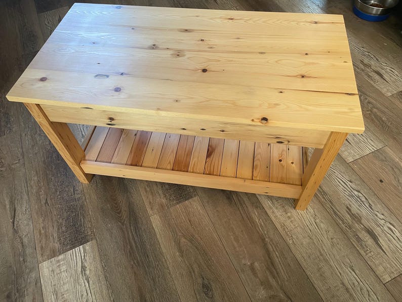 May include: A rectangular coffee table constructed from light-colored natural wood with visible grain patterns and dark knots. The table features a solid flat top surface and a lower slatted shelf supported by four sturdy wooden legs.