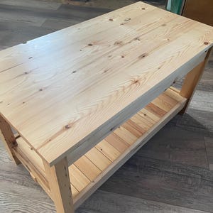 May include: A rectangular wooden coffee table constructed from light-colored pine wood with a visible grain pattern and natural knots. The table features a smooth flat top surface and a lower slatted shelf supported by four sturdy vertical legs.