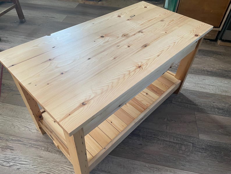 May include: A rectangular wooden coffee table constructed from light-colored pine wood with a visible grain pattern and natural knots. The table features a smooth flat top surface and a lower slatted shelf supported by four sturdy vertical legs.