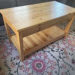 May include: A rectangular coffee table constructed from light-toned wood with a visible grain and natural knots. The table features a solid flat top surface and a lower slatted shelf supported by four sturdy, straight legs at each corner.