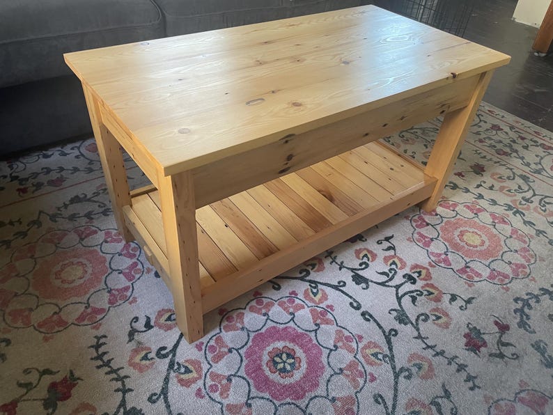 May include: A rectangular coffee table constructed from light-toned wood with a visible grain and natural knots. The table features a solid flat top surface and a lower slatted shelf supported by four sturdy, straight legs at each corner.