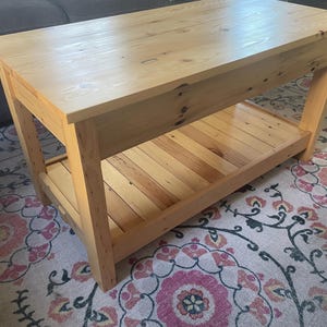 May include: A rectangular coffee table constructed from light-colored natural wood with a smooth top surface and a slatted lower shelf. The table features four sturdy legs and visible wood grain patterns throughout the entire structure.
