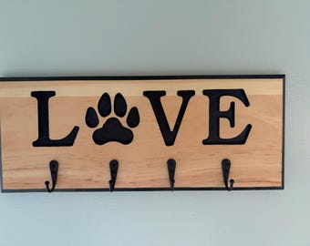 May include: A rectangular wooden wall hook rack featuring the word LOVE in black lettering, with a black paw print replacing the letter O. The natural light-colored wood board has a black border and four black metal hooks attached at the bottom.