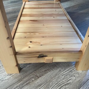 May include: A light-colored wooden table featuring a slatted lower shelf constructed from horizontal planks. The table has four thick, square-cut legs and a frame with visible wood grain, knots, and a small central wooden block detail on the front.