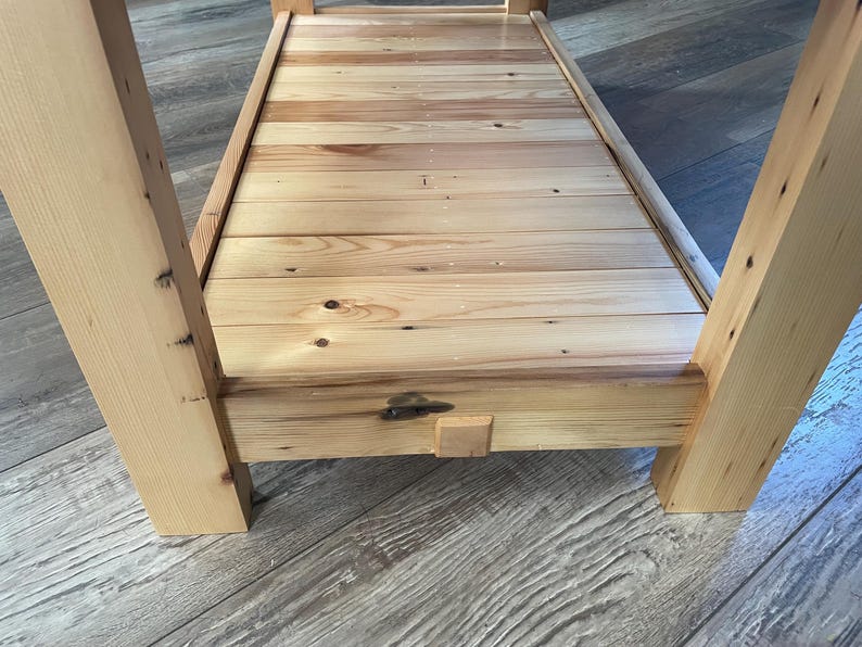May include: A light-colored wooden table featuring a slatted lower shelf constructed from horizontal planks. The table has four thick, square-cut legs and a frame with visible wood grain, knots, and a small central wooden block detail on the front.