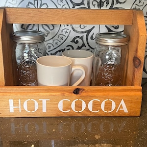 May include: A light brown wooden caddy with a handle featuring the white text HOT COCOA on the front. Inside the caddy are two clear glass mason jars with silver metal lids and two plain white ceramic mugs arranged in a single row.