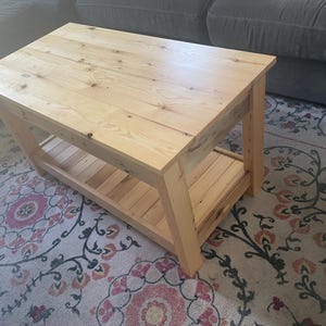 May include: A rectangular coffee table constructed from light-colored natural wood with visible grain patterns and knots. The table features a solid flat top surface supported by four sturdy legs and includes a lower slatted shelf for storage.