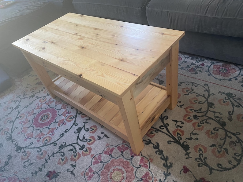 May include: A rectangular coffee table constructed from light-colored natural wood with visible grain patterns and knots. The table features a solid flat top surface supported by four sturdy legs and includes a lower slatted shelf for storage.