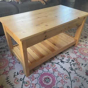 May include: A rectangular light-colored wooden coffee table featuring a smooth top surface with visible wood grain and knots. The table is supported by four sturdy legs and includes a slatted lower shelf for additional storage space.