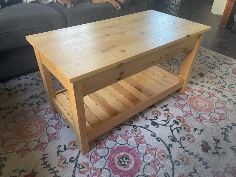 May include: A rectangular light-colored wooden coffee table featuring a smooth top surface with visible wood grain and knots. The table is supported by four sturdy legs and includes a slatted lower shelf for additional storage space.