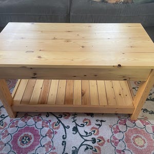 May include: A rectangular light-colored wooden coffee table featuring a smooth top surface with visible wood grain and knots. The table is constructed with four sturdy legs and includes a slatted lower shelf for storage, showcasing a natural finish.