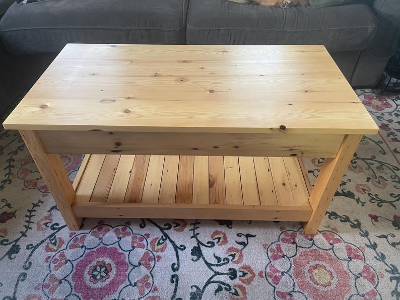 May include: A rectangular light-colored wooden coffee table featuring a smooth top surface with visible wood grain and knots. The table is constructed with four sturdy legs and includes a slatted lower shelf for storage, showcasing a natural finish.