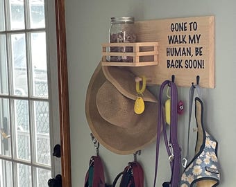 May include: A light-colored wooden wall-mounted organizer featuring a small shelf with a glass jar, three black hooks, and the black text GONE TO WALK MY HUMAN, BE BACK SOON!. A tan sun hat, a purple leash, and a daisy-patterned pet harness hang below.