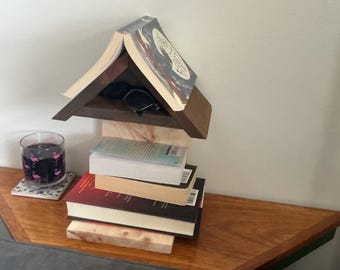 May include: A wooden book stand constructed from stacked books and dark wood pieces, forming a house-like structure. The top book is open like a roof, while the base consists of horizontal books. A glass with a flamingo pattern sits nearby.