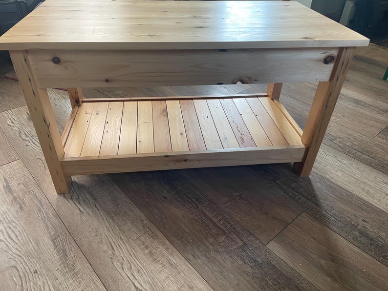 May include: A rectangular wooden table featuring a smooth, light-toned natural wood grain finish. The table has four sturdy legs and a lower slatted shelf for storage. The construction displays visible knots and wood grain patterns throughout the surface.