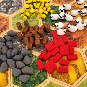 Op de afbeelding: Een bordspel met diverse spelstukken. De stukken omvatten gele tarwebundels, witte schapen met zwarte gezichten, bruine boomstammen, grijze stenen en rode bakstenen. Het spelbord is een zeshoekig raster met terreintegels.