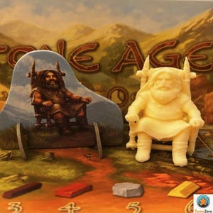 Può includere: Un gioco da tavolo con il titolo "Stone Age" visibile sullo sfondo. Il tabellone presenta una figura seduta su una sedia e una statuina 3D di un uomo seduto. I pezzi del gioco includono blocchi colorati marroni, rossi, grigi e dorati.