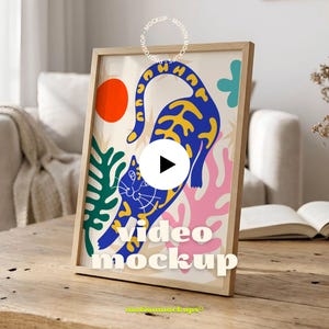 May include: A framed art print featuring a blue cat with yellow accents, a red circle, and abstract shapes in green and pink. The frame is light wood, and the print has the words "Video Mockup".