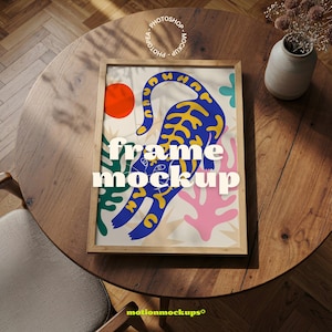 May include: A framed art print featuring a blue tiger with yellow accents, an orange sun, and colorful abstract shapes. The text "frame mockup" is displayed on the artwork. The frame is light brown and sits on a wooden table.