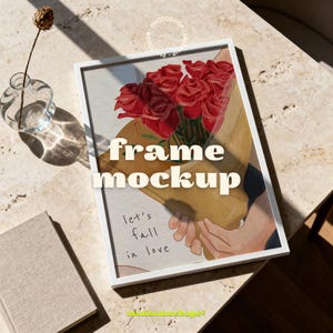 May include: White framed art print with a bouquet of red roses, the text "frame mockup" and "let's fall in love". The print is on a marble surface, with a small vase and a book.