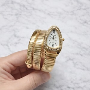 Op de afbeelding: Goudkleurig polshorloge met een uniek ontwerp. Het horloge heeft een driehoekige wijzerplaat met een witte wijzerplaat, Romeinse cijfers en een bezel versierd met kleine kristallen. De band is een flexibel, opgerold ontwerp. Het horloge is ongeveer 18 cm lang.