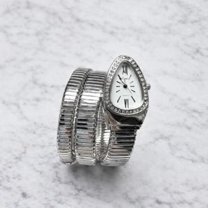 Puede incluir: Un reloj de pulsera plateado con un diseño único en forma de serpiente. La esfera es blanca con manecillas negras y números romanos. El bisel está incrustado con pequeños cristales transparentes. La correa se enrolla varias veces alrededor de la muñeca.