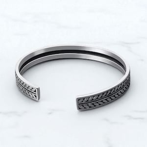 May include: Silver cuff bracelet with an open design. The bracelet has a detailed leaf pattern on the outer edges and a sleek inner band. The jewellery is displayed on a white marble surface.